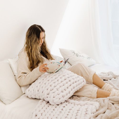 A woman with long wavy hair sits on a bed covered in white bedding, reading a book, with a chunky knitted blanket on her lap in a bright, cosy room—Bring Hygge into Your Bedroom for ultimate comfort and warmth.