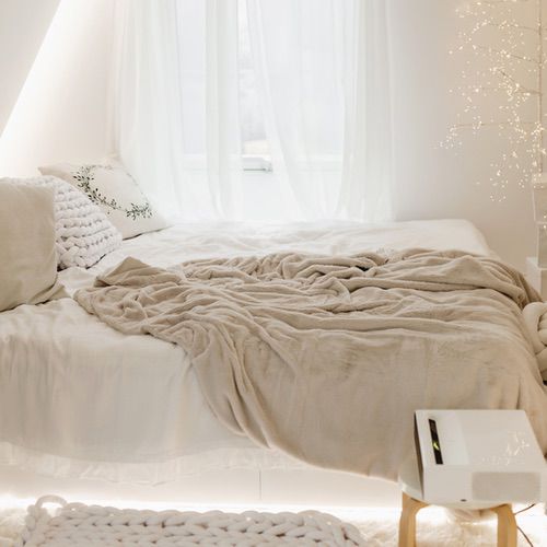 A cosy, softly lit bedroom with a neatly made bed covered in cream-coloured blankets and pillows. Sheer white curtains let in natural light—bring hygge into your bedroom with a decorative tree adorned with fairy lights beside the bed.