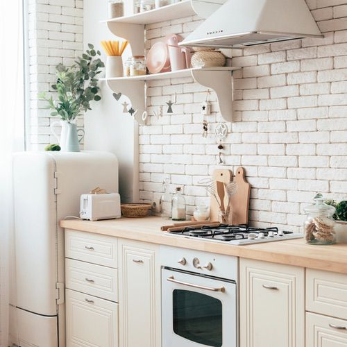 A bright, cosy kitchen with white cupboards, a vintage fridge, a gas cooker, wooden worktops, and shelves with crockery and plants against a white brick wall. Bring Hygge into Your Kitchen as natural light streams through the nearby window.