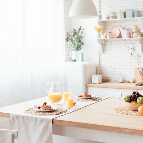 Bring Hygge into Your Kitchen with this bright, airy space—a wooden table set for breakfast, pancakes and fresh juice under sunlight streaming through sheer curtains. Cosy shelves display charming crockery and kitchenware.