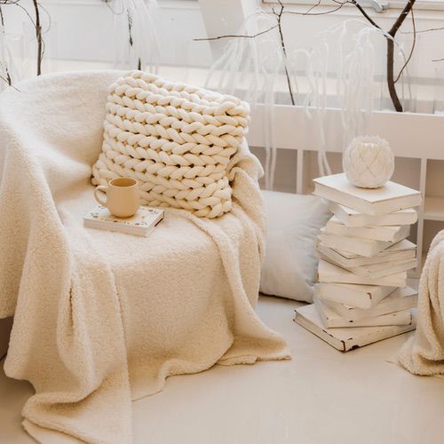 Bring Hygge into Your Living Room with this cosy beige chair, draped in a fluffy blanket and chunky knit cushion, beside whitewashed books topped with a candle; a mug and book invite you to relax.