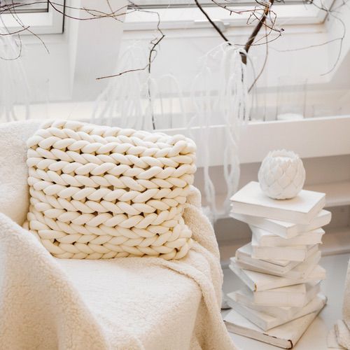 Bring Hygge into Your Living Room with a cosy chair featuring a chunky knit white cushion and soft blanket, nestled by a stack of white books and dried branches in a bright, minimalist space with large windows.