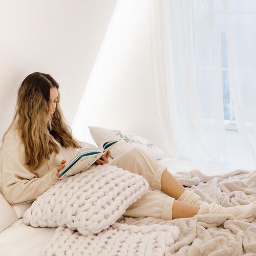A woman with long hair sits on a bed by a window, wearing cosy loungewear and socks. She enjoys hygge activities like reading a book, surrounded by soft, chunky knit blankets in a bright, peaceful room.
