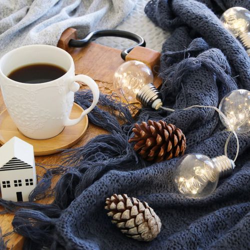 A cosy scene for Hygge for Beginners: a cup of black coffee on a wooden tray, dark blue knitted blanket, pinecones, glowing fairy lights with large bulbs, and a small ceramic house decoration.