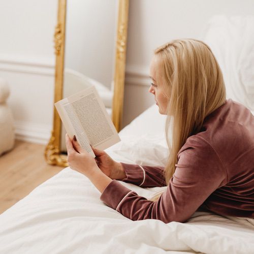 A woman with long blonde hair lies on a bed in a cosy, hyggeable room, wearing a mauve pyjama top and reading a book. A large ornate mirror leans against the wall in the background.