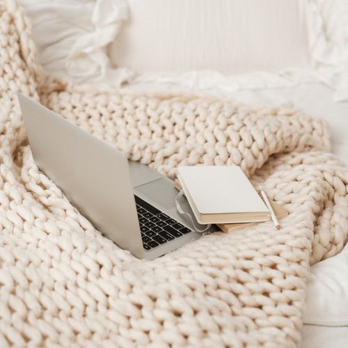 A silver laptop, a closed notebook, and a pencil rest on a chunky knit cream blanket atop a cosy white bed with fluffy pillows—perfect for reviewing the Hyggeable Terms and Conditions in comfort.
