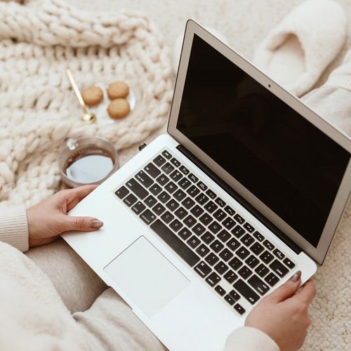 A person in cosy clothing sits on a knitted blanket with a laptop on their lap, browsing Hyggeable website disclaimers, next to a cup of coffee and a small plate of biscuits on a soft beige surface.