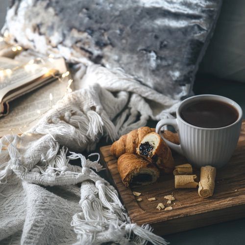 A cosy scene with a cup of hot chocolate and pastries on a wooden tray, set on a bed with a soft textured blanket, an open book, and fairy lights in the background—everything that shows what hygge isn't: cold or uninviting.