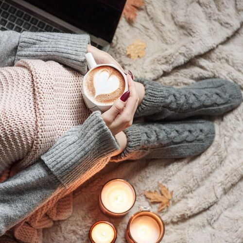 A person in cosy grey knit socks and a pink jumper sits on a soft blanket, holding a cup of latte with heart-shaped foam art. Three lit candles and autumn leaves are nearby, showing what hygge isn't—loud, hectic, or uncomfortable, but warm and relaxing.
