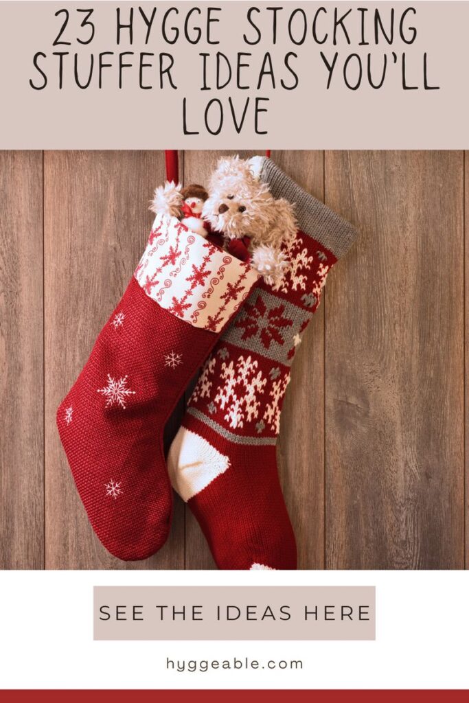 Two red and white holiday stockings with snowflake patterns hang on a wooden wall. One holds a small soft teddy bear. Text above reads, 23 Hygge Stocking Stuffer Ideas You'll Love—perfect cosy inspiration for your holiday season.