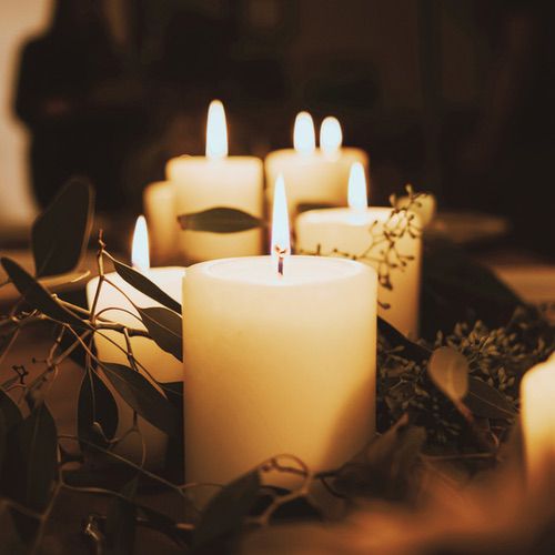 A group of lit white pillar candles for hygge, surrounded by green leaves and foliage, creates a warm, cosy, and inviting atmosphere.