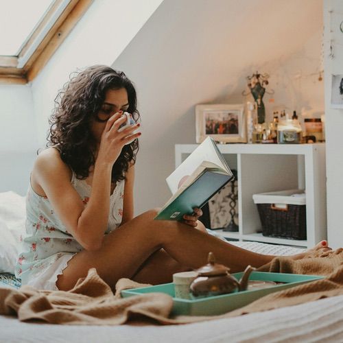 A woman with curly hair sits on a bed by a window, wearing pyjamas, drinking from a cup and reading a book in her cosy reading nook. A tray with a teapot and cup rests in front of her, filling the softly lit room with warmth.
