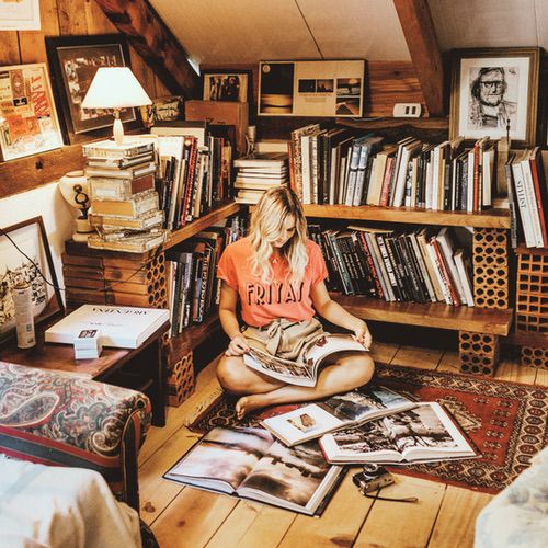 A woman sits cross-legged on a rug in a cosy reading nook within an attic library, reading large books spread around her. Bookshelves line the walls, and the warm lighting creates a relaxed, inviting atmosphere.