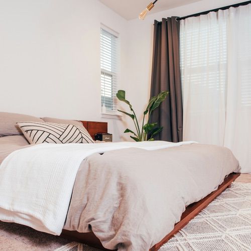A modern hygge bedroom with a low wooden bed, beige bedding, a black and white cushion, white blanket, potted plant beside the bed, white walls, large windows with airy curtains, and a patterned rug.