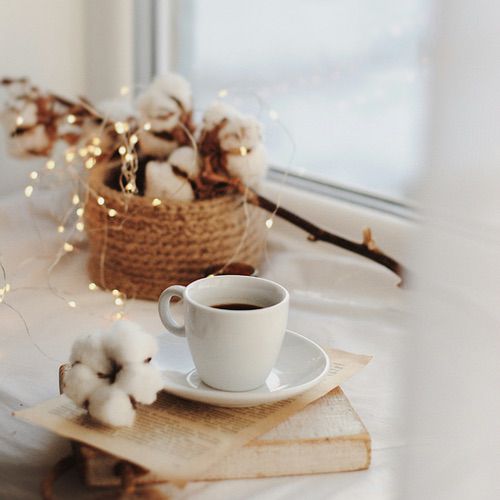 A white cup of coffee sits on a saucer atop an old book, creating a cosy hygge drink ritual, with cotton flowers and a woven basket decorated with fairy lights in the background near a sunlit window.