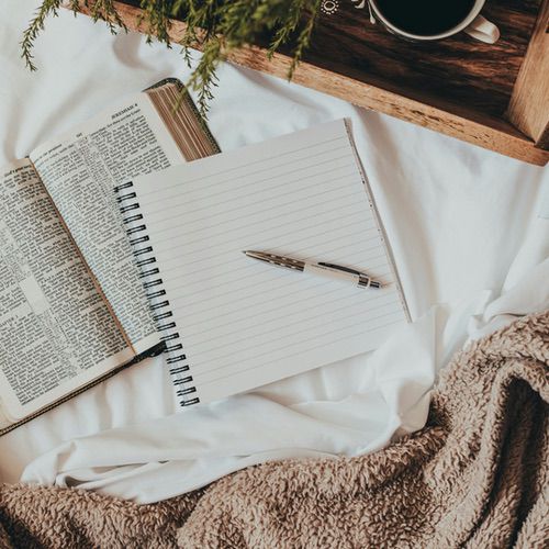 A spiral notebook ideal for hygge journaling rests on a bed beside an open book, a brown blanket, and a wooden tray with coffee and greenery.