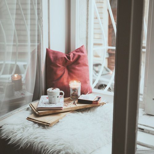 A cosy window seat with a red cushion, a fur throw, a lit candle, hot chocolate with whipped cream, books, and envelopes—perfect for hygge journalling—next to a window with white bars and soft natural light.
