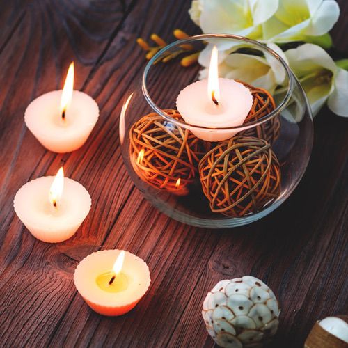 Four lit tealight candles are arranged on a wooden surface next to a glass bowl of decorative wicker balls. White flowers and another decorative ball sit nearby, offering inspiration for cosy hygge routine ideas and creating a relaxing atmosphere.