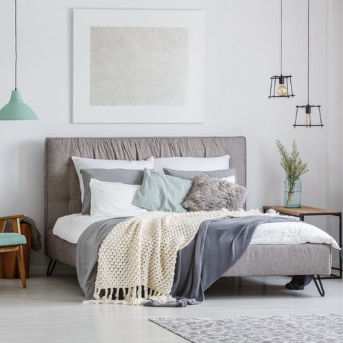 Modern bedroom with a grey upholstered bed, cosy pillows, and a textured blanket invites cosy Hygge bedtime rituals. Two bedside tables with lamps, greenery in a vase, and framed abstract artwork complete the serene, neutral space.