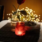 A red rose in a glass vase, a lit red candle, and a bowl of chocolates on a table, set against a backdrop of cosy mood lighting.