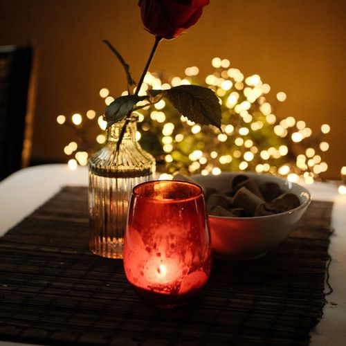 A red rose in a glass vase, a lit red candle, and a bowl of chocolates on a table, set against a backdrop of cosy mood lighting.