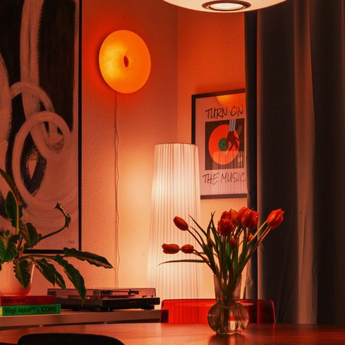 A cosy room interior with warm mood lighting from an orange wall light and a tall floor lamp, featuring a vase of red tulips on a table and framed art on the walls.