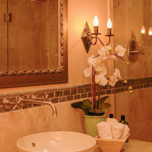 A bathroom vanity with a white vessel basin, a wall-mounted tap, a potted white orchid, and a basket of rolled flannels, all bathed in soft hygge bathroom lighting.