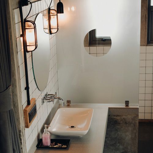 A modern bathroom vanity featuring a white rectangular basin, a round mirror, and warm hygge bathroom lighting mounted above a tiled wall.