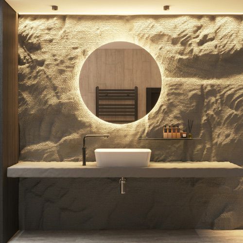 A backlit circular mirror hangs above a white rectangular basin on a stone shelf against a textured rock wall, creating warm hygge bathroom lighting.