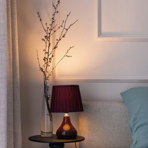 A small bedside table lamp with a dark red pleated shade next to a tall glass vase containing bare branches, perfect for hygge bedroom lighting ideas.