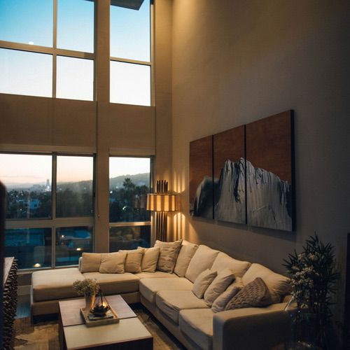 A spacious lounge featuring a large beige corner sofa and a mountain peak triptych, accented by hygge lighting ideas for the lounge and tall windows overlooking a city at dusk.