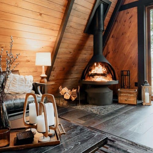A modern A-frame cabin interior featuring a crackling wood-burning fireplace, a leather sofa, and a wooden coffee table with pillar candles that offer cosy hygge lighting ideas for the lounge.