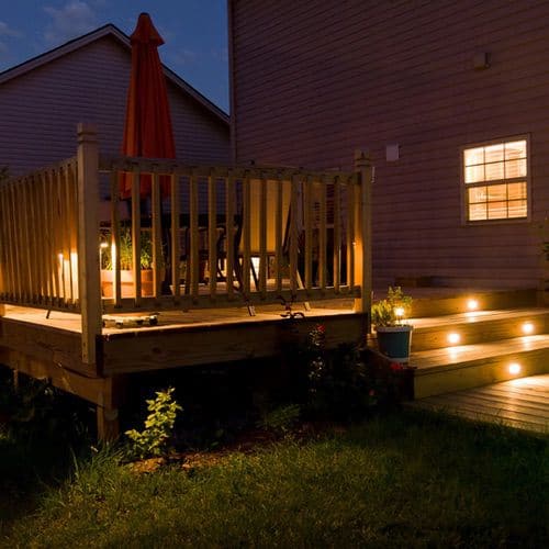 A wooden deck at dusk showcases hygge outdoor lighting ideas with a patio table, an orange parasol, and built-in step lights illuminating the stairs.