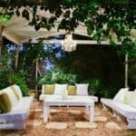Outdoor seating area with white sofas and a white coffee table under a vine-covered pergola with a hanging chandelier for cosy hygge outdoor lighting.