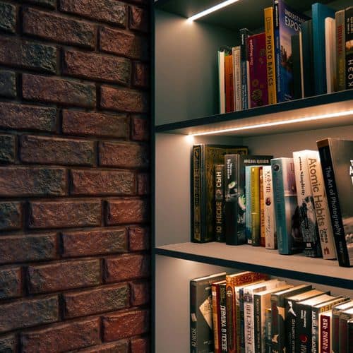 A bookcase with three tiers of books illuminated by built-in LED lighting, positioned next to a dark brick wall. Lighting Bookshelves ideas