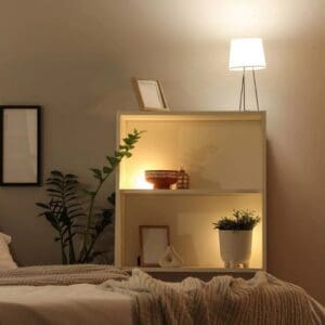 A white bookcase in a bedroom with lighting on top, containing decor items and a potted plant, with another plant to the side. Lighting Bookshelves ideas